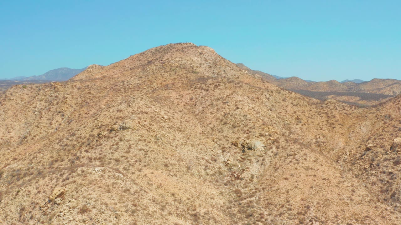 Mid-clip frame of a rocky desert slope near Dreams Los Cabos, with shallow gullies, sparse shrubs, distant brown hills, clear sky, and no visible people.