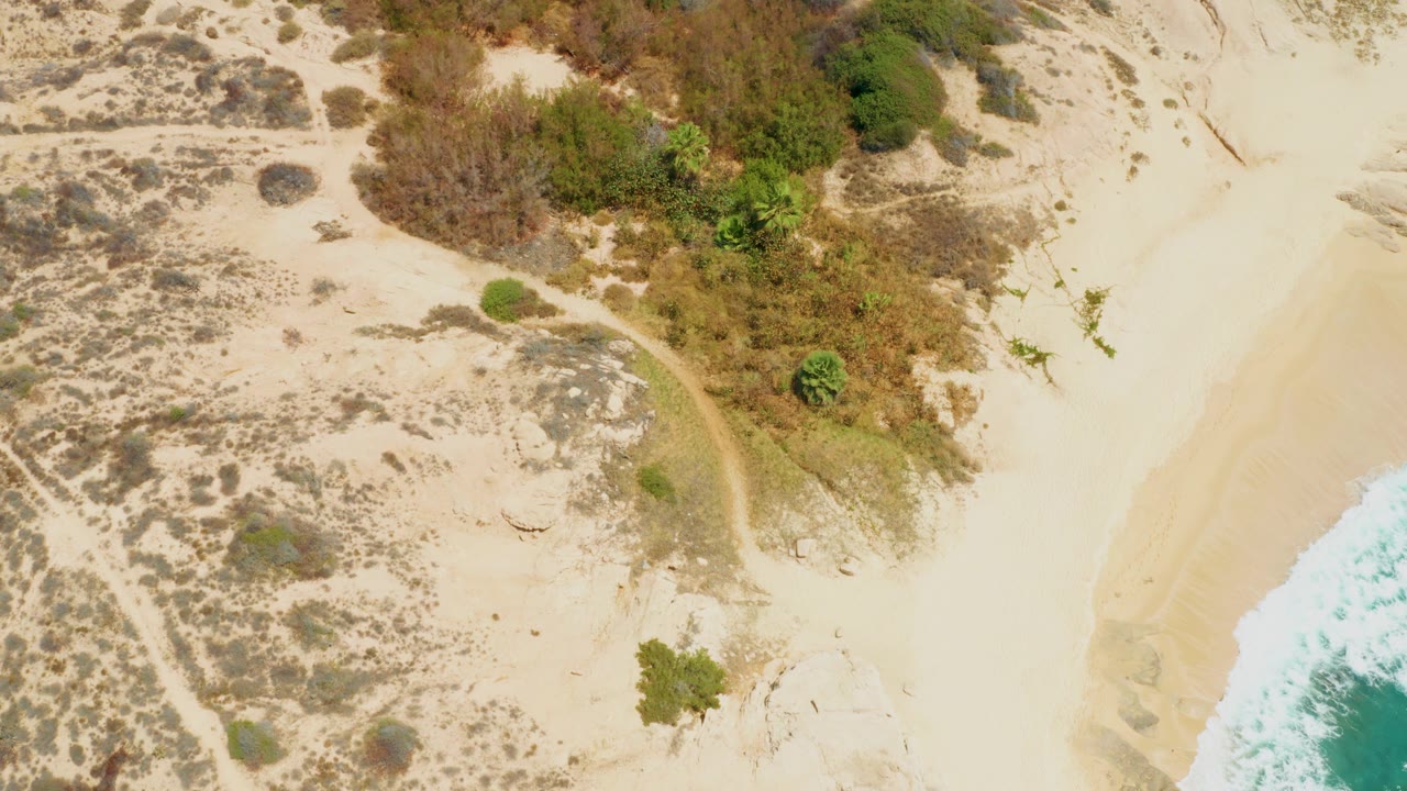Drone-height frame at Dreams Los Cabos with scrub-covered dunes on the left, smoother open sand toward the beach, surf at the right border, and no visible people.