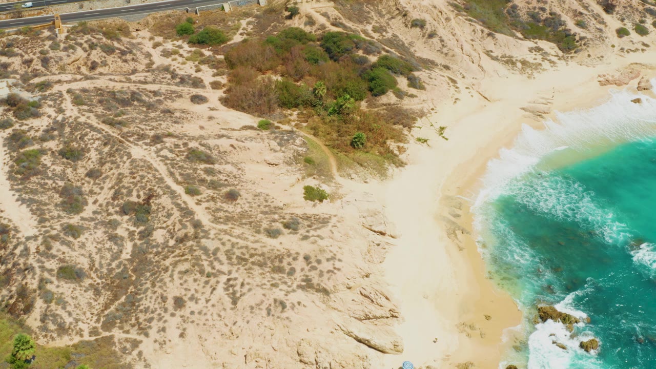 Aerial frame over Dreams Los Cabos showing pale dunes with scrub, a curving beach, turquoise water, white foam, a road near the top edge, and a tiny person on the sand.