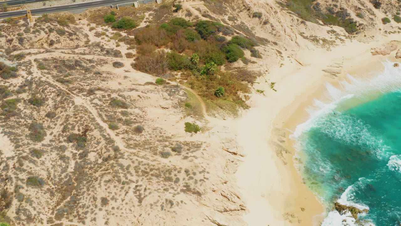 Aerial frame over Dreams Los Cabos showing the upper road, dry dune vegetation, a smooth sandy beach, blue-green ocean water, and a very small person on the shoreline sand.