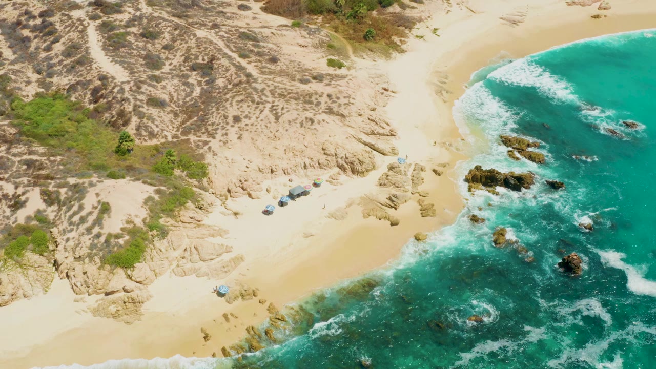 Aerial frame over Dreams Los Cabos showing pale sand, scrub-covered dunes, turquoise water, rock outcrops, small shade structures, and no visible people.