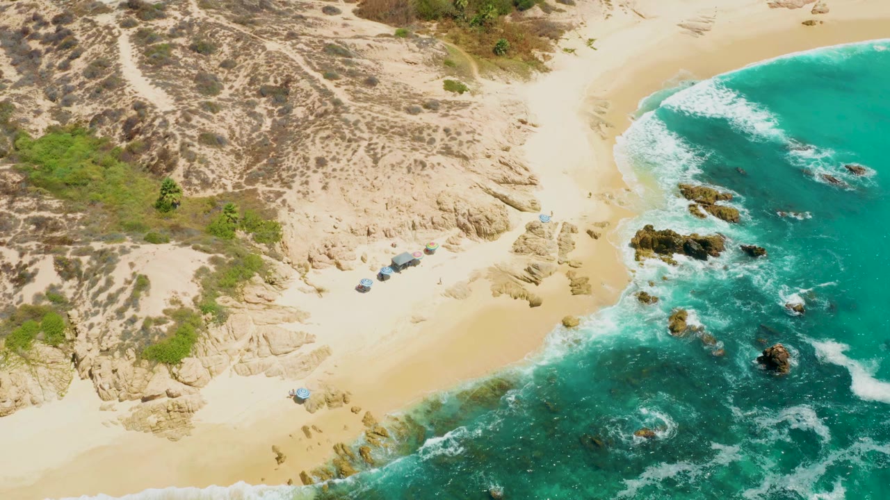 Overhead coastal view with dunes, sand, beach structures, rocks, and teal water.