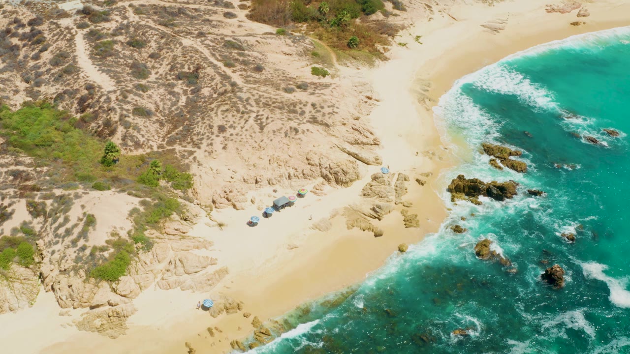 Overhead frame from Dreams Los Cabos showing a curved sandy shoreline, bright turquoise water, foamy waves, rocks near the waterline, scrub dunes, and no visible people.