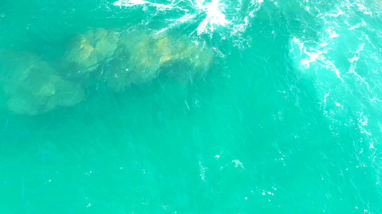 Overhead view of turquoise ocean water at Dreams Los Cabos with submerged rocks near the upper left and white surface foam near the top, no visible people.