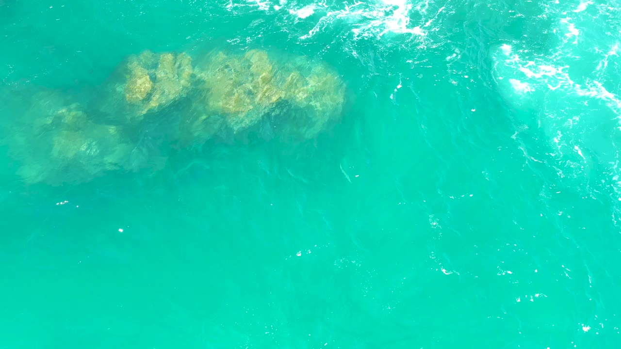 A tan rocky patch sits beneath turquoise ocean water near the upper right of the overhead frame, with white foam entering from the top edge, no visible people.