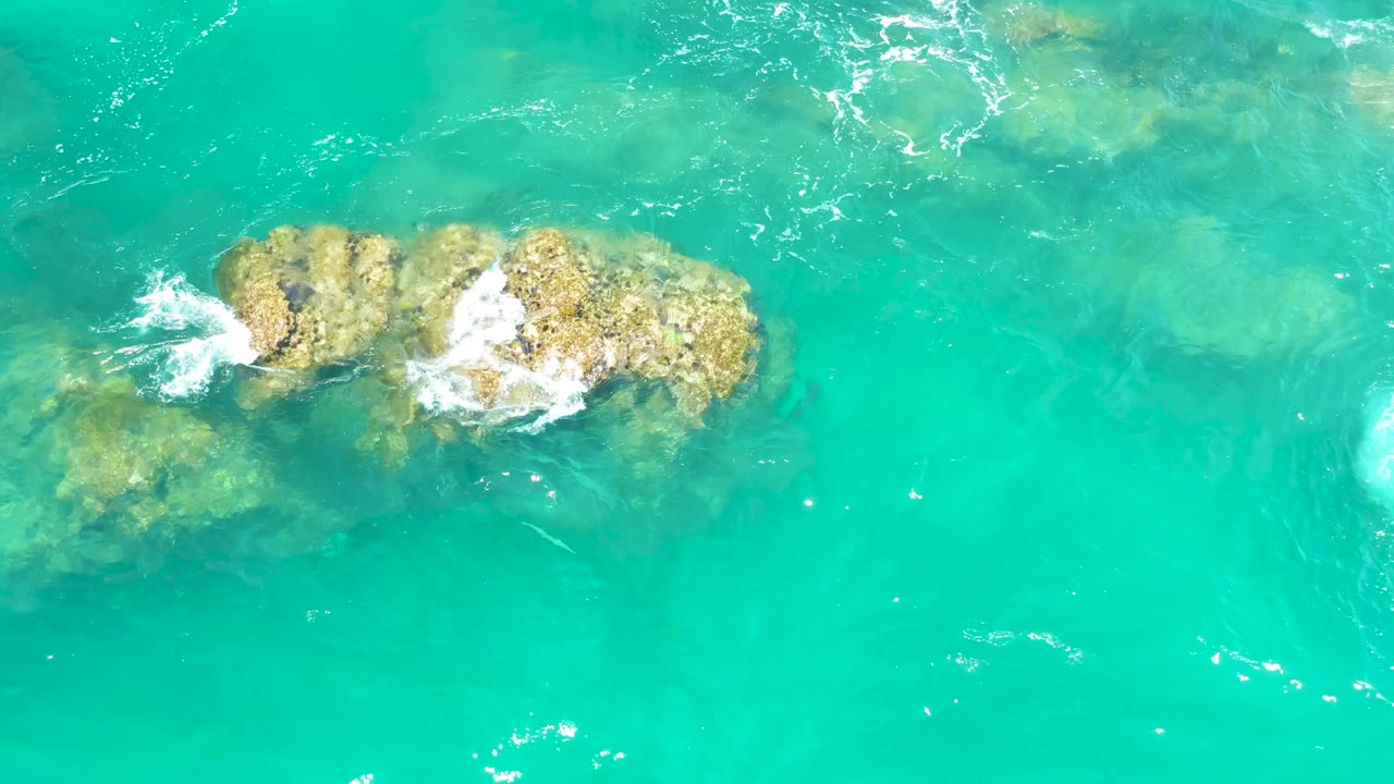 At Dreams Los Cabos in Baja California Sur, white foam spreads farther across the main rock group while additional submerged rocks sit below in clear turquoise water, with no visible people.
