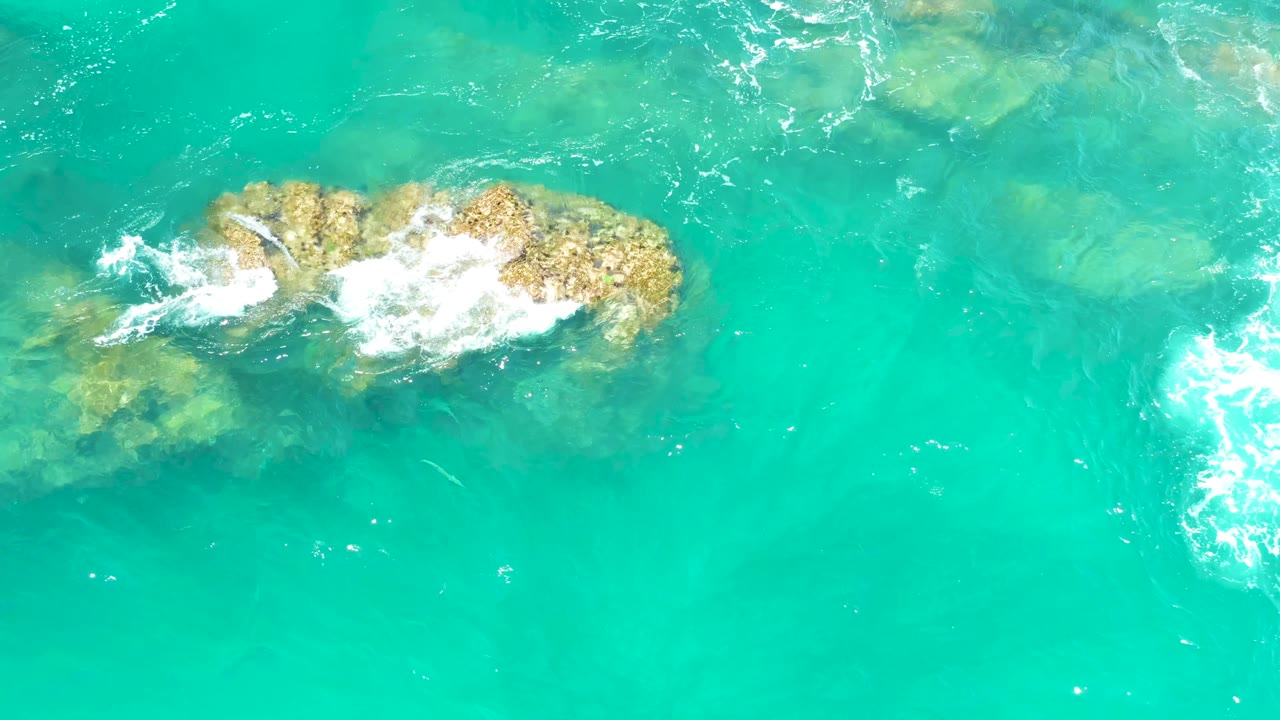 At Dreams Los Cabos in Baja California Sur, the foam concentrates on the right half of the exposed rock while pale submerged formations remain visible through the turquoise water, with no visible people.