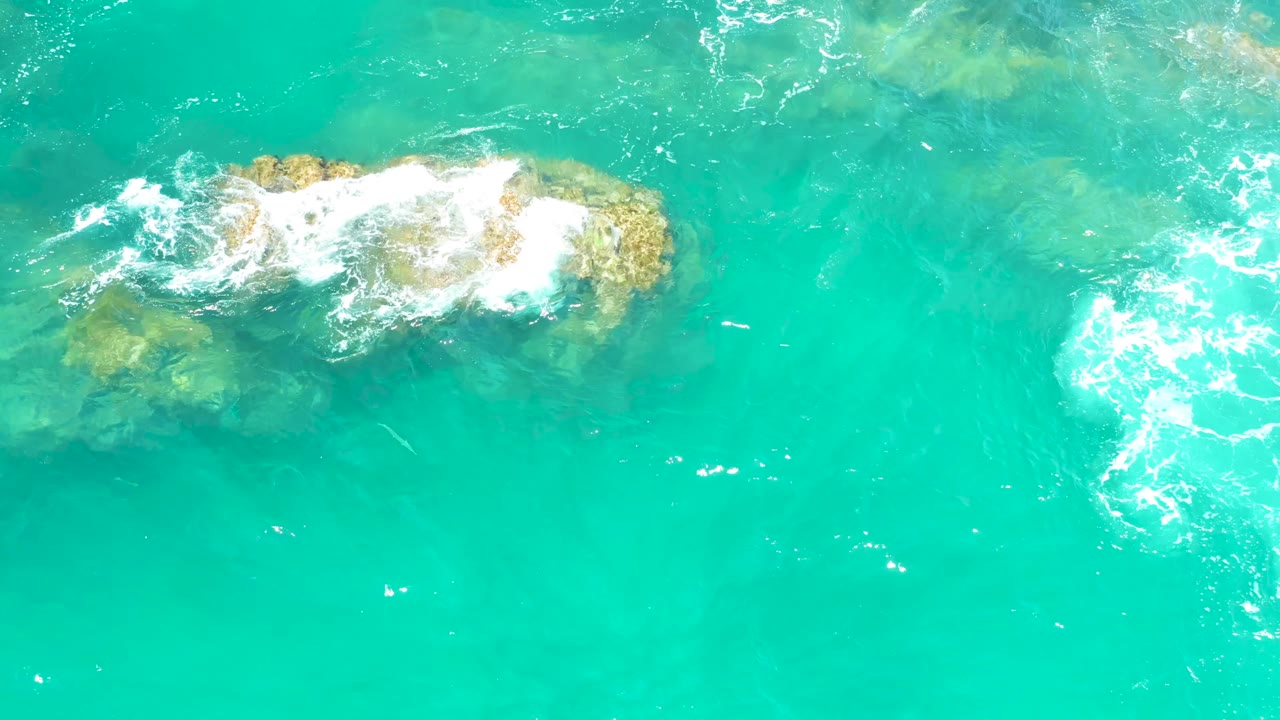 At Dreams Los Cabos in Baja California Sur, a wider sheet of foam moves across the rock line while the surrounding blue-green water remains clear enough to show darker seabed shapes, with no visible people.