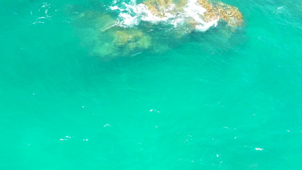 Aerial frame from Dreams Los Cabos showing clear turquoise water, a partially submerged rocky outcrop near the top, and fresh white foam crossing the rocks, with no visible people.