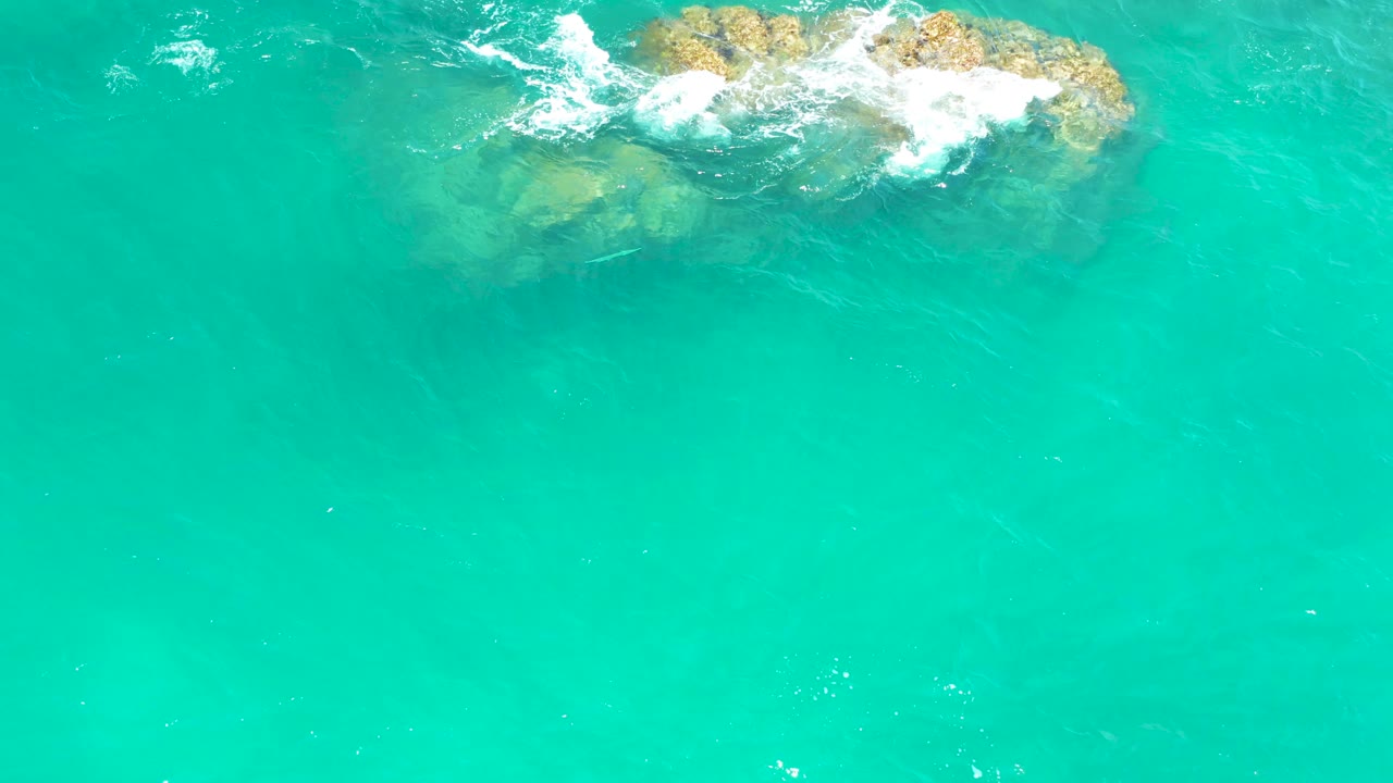 Overhead Dreams Los Cabos water detail with the rocky outcrop shifted toward the upper right and white surf curling over it, with no visible people.