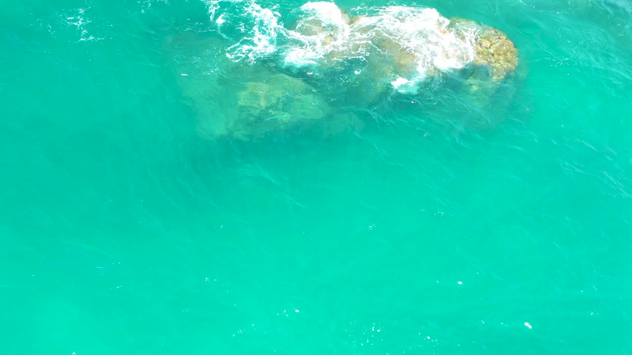 Aerial view at Dreams Los Cabos with foamy white water spreading over a submerged rock cluster on the left side of a bright turquoise frame, with no visible people.