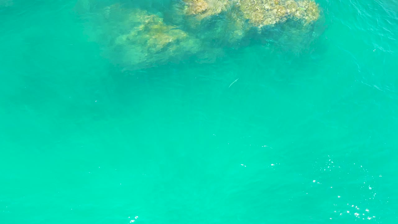 Submerged tan rocks sit beneath clear turquoise water at Dreams Los Cabos, with ripples crossing the surface and no visible people.