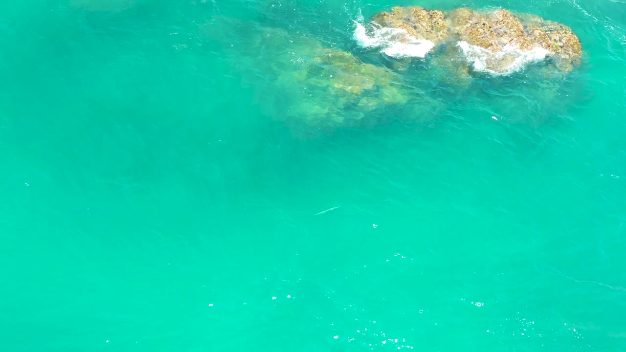 Aerial frame from Dreams Los Cabos with the rock cluster still near the upper edge, white foam thinning around it, and no visible people.