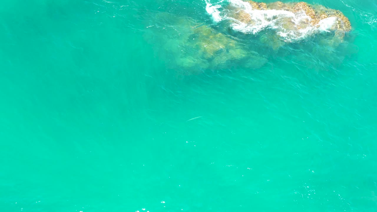 Aerial view over Dreams Los Cabos water where the rocks sit high in frame, surf breaks in small white patches, and open turquoise water fills the foreground with no visible people.
