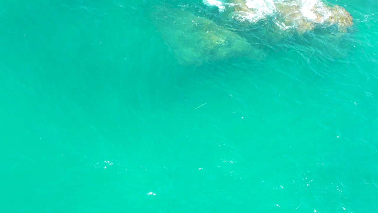 Dreams Los Cabos aerial frame with the rocks shifted toward the upper left, a thin line of white foam, broad rippled water below, and no visible people.