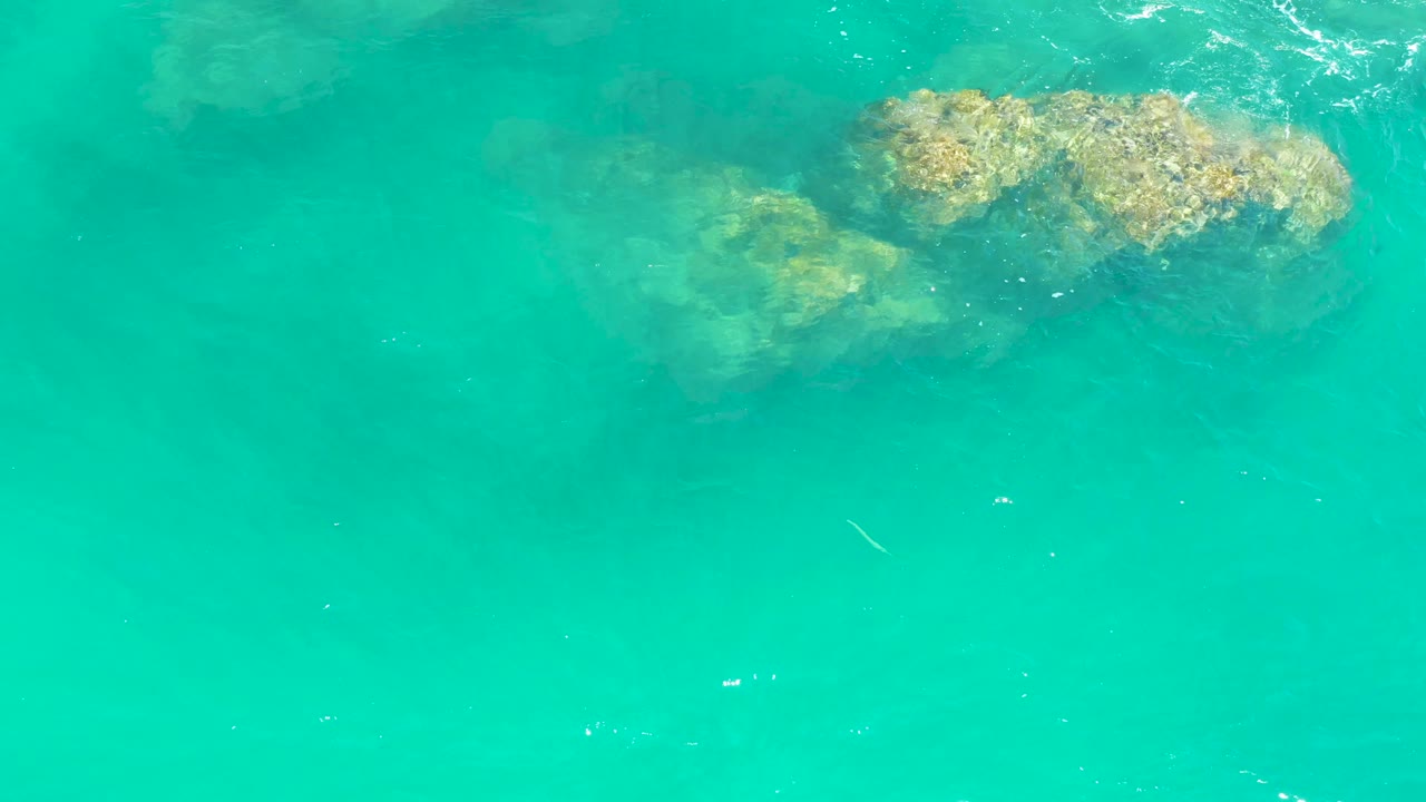 Clear turquoise ocean water shows the rocky shelf just below the surface at Dreams Los Cabos, with light foam near the upper edge and no visible people.