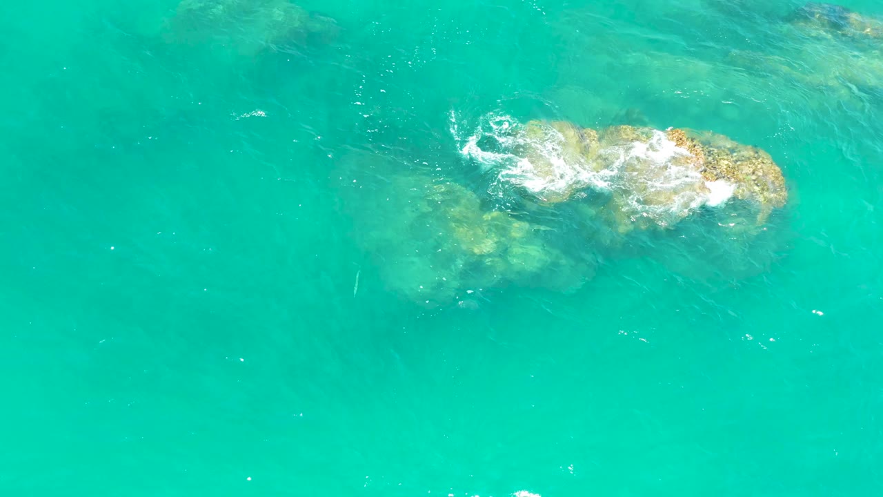 Aerial view at Dreams Los Cabos, Mexico, where white foam moves across the top of a shallow rock patch in bright turquoise water, with no visible people.