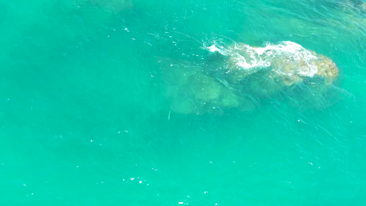 Top-down view of clear turquoise water at Dreams Los Cabos, Mexico, with a low rock formation under the surface and a small wash of foam, with no visible people.