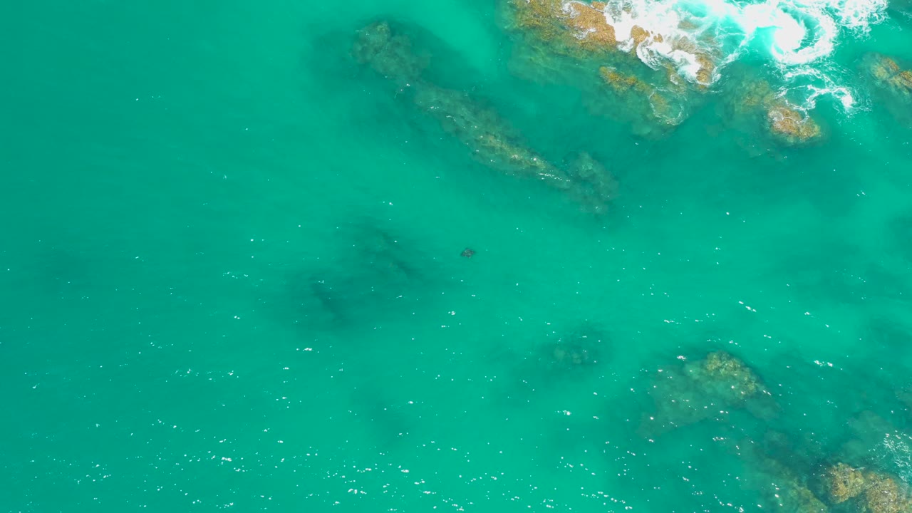 Top-down ocean view at Dreams Los Cabos, Mexico, where white foam recedes near the upper right and shallow rocks remain visible through turquoise water, with no visible people.