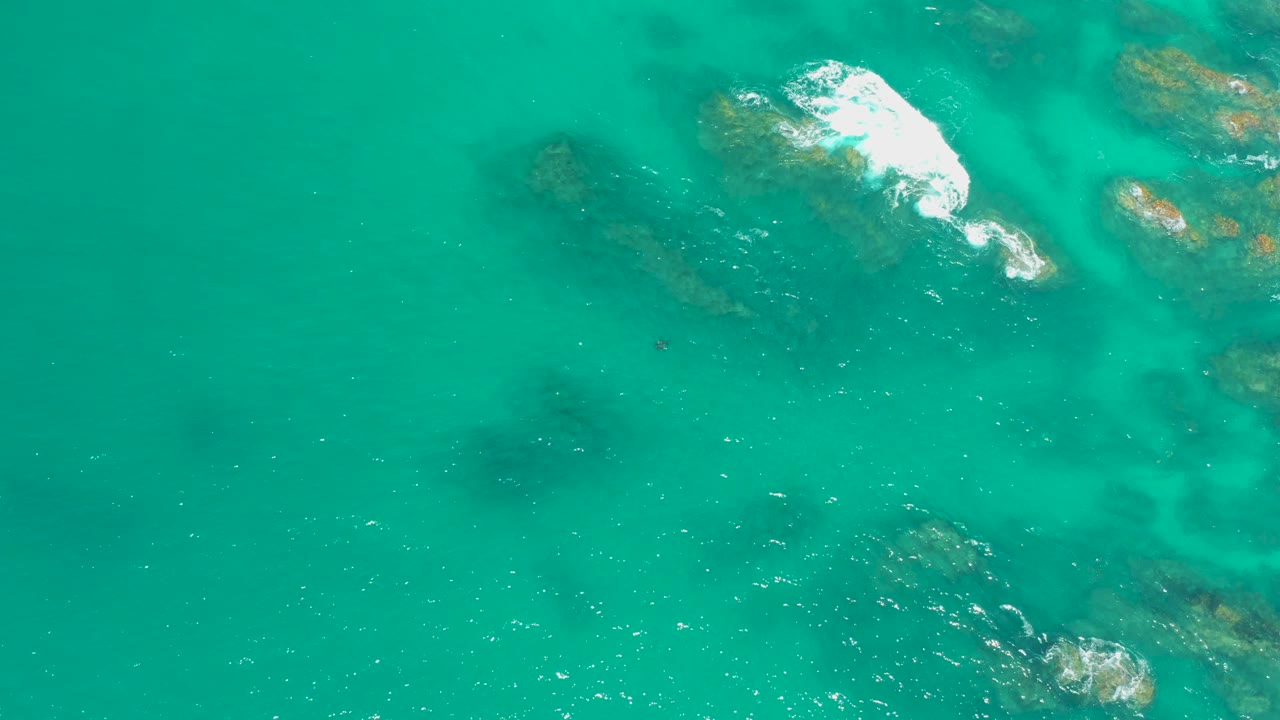 Overhead view at Dreams Los Cabos showing turquoise water, submerged rocky seabed, a small patch of white wave foam near the upper frame, and no visible people.