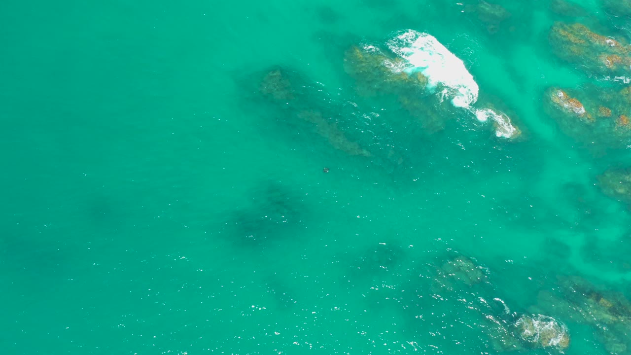Overhead Dreams Los Cabos frame with clear teal water, dark submerged rocks, white surf breaking in the upper right, and no visible people.