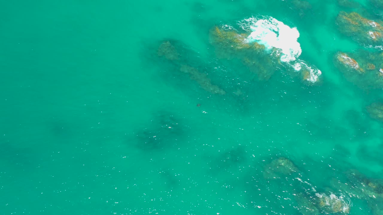 Aerial view over Dreams Los Cabos water with scattered rocks below the turquoise surface, a white foam patch near the top edge, and no visible people.