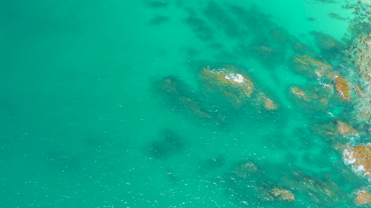 Aerial view at Dreams Los Cabos with open turquoise water on the left, underwater rocks toward the right, small foam, and no visible people.