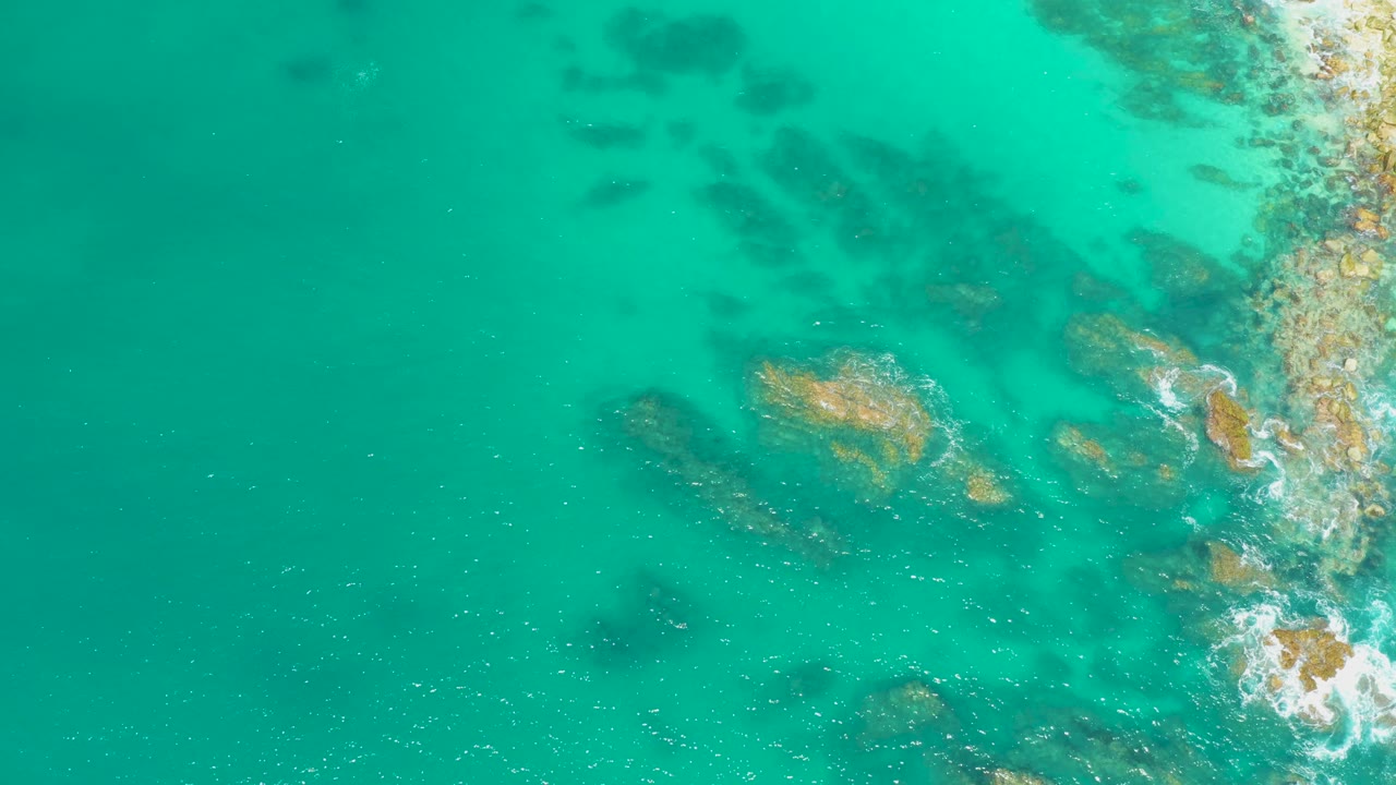 Clear green-blue water at Dreams Los Cabos reveals darker submerged rock patches, tan exposed rocks, and a narrow rocky edge with no visible people.