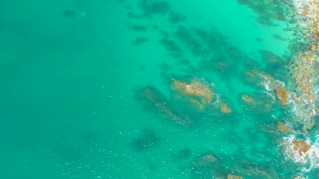 Aerial frame from Dreams Los Cabos with turquoise water, scattered submerged rocks, small white breaks against the right-side shoreline, and no visible people.