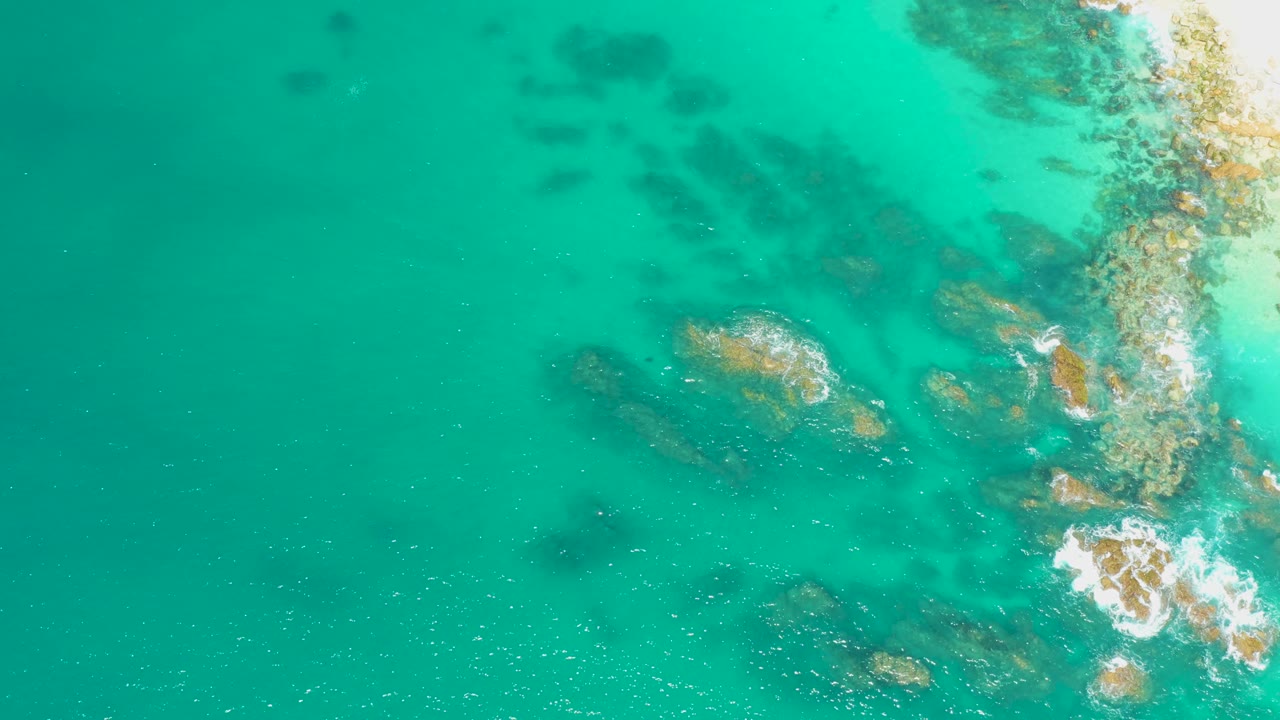 Overhead view of the Dreams Los Cabos shoreline where turquoise water surrounds brown rocks and a brighter foam cluster forms near the lower right, with no visible people.