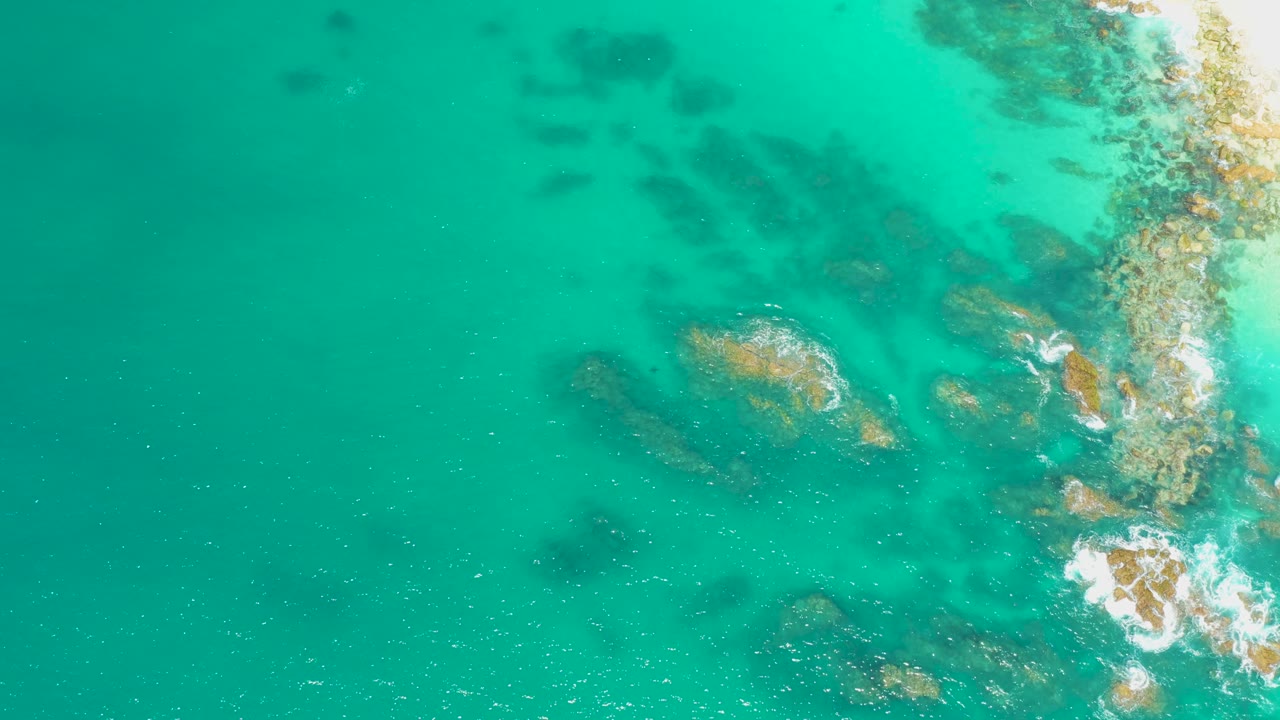 Aerial view at Dreams Los Cabos with open turquoise water on the left, submerged rocks through the center, and small foam lines against the rocky shore, with no visible people.