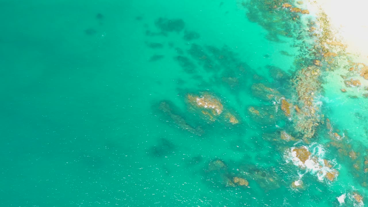 Overhead view at Dreams Los Cabos of turquoise water, submerged rocky patches, and white foam breaking near the right side, with no visible people.