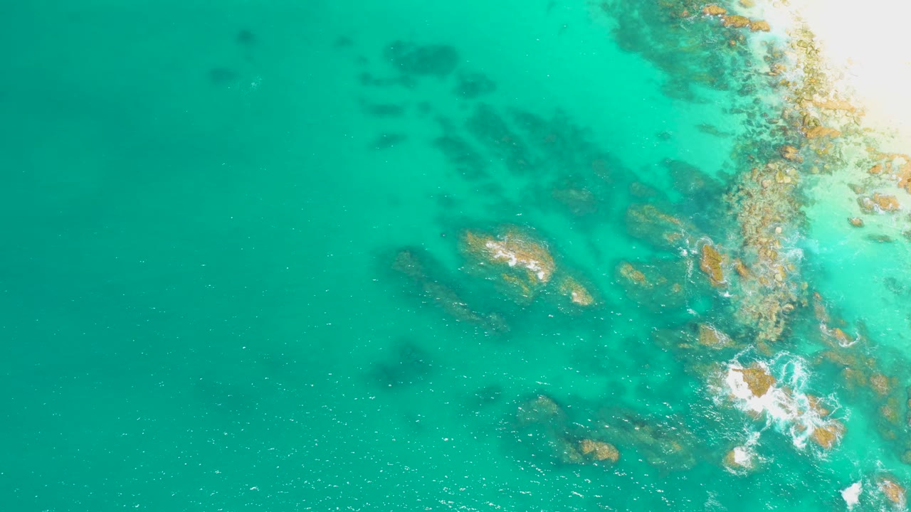Overhead Dreams Los Cabos shoreline detail with turquoise water, scattered submerged rocks, and small white wave breaks shifted along the right side, with no visible people.