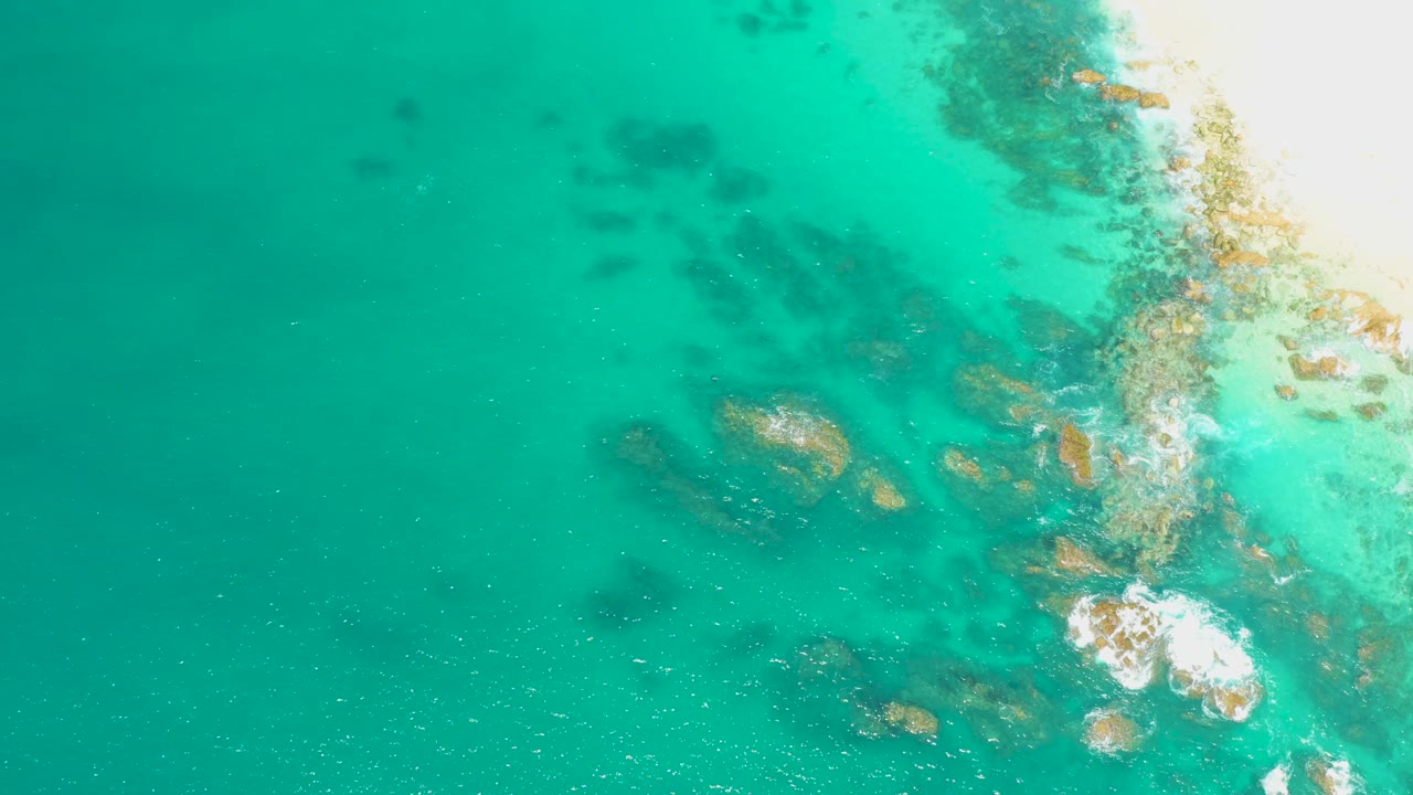 High overhead view at Dreams Los Cabos showing turquoise shallow water, submerged rocks, white wave foam near the bright shoreline edge, and no visible people.