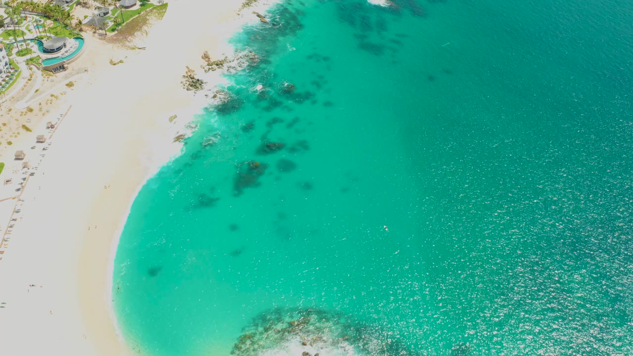 Opening aerial frame over Dreams Los Cabos with pale sand on the left, clear turquoise water on the right, dark underwater rock patches near shore, and no visible people.