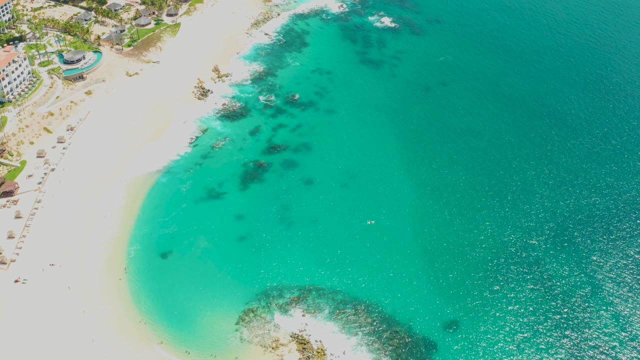Aerial view at Dreams Los Cabos with the curved white shoreline along the left, pale turquoise shallows near the sand, darker rock patches under the water, and no visible people.