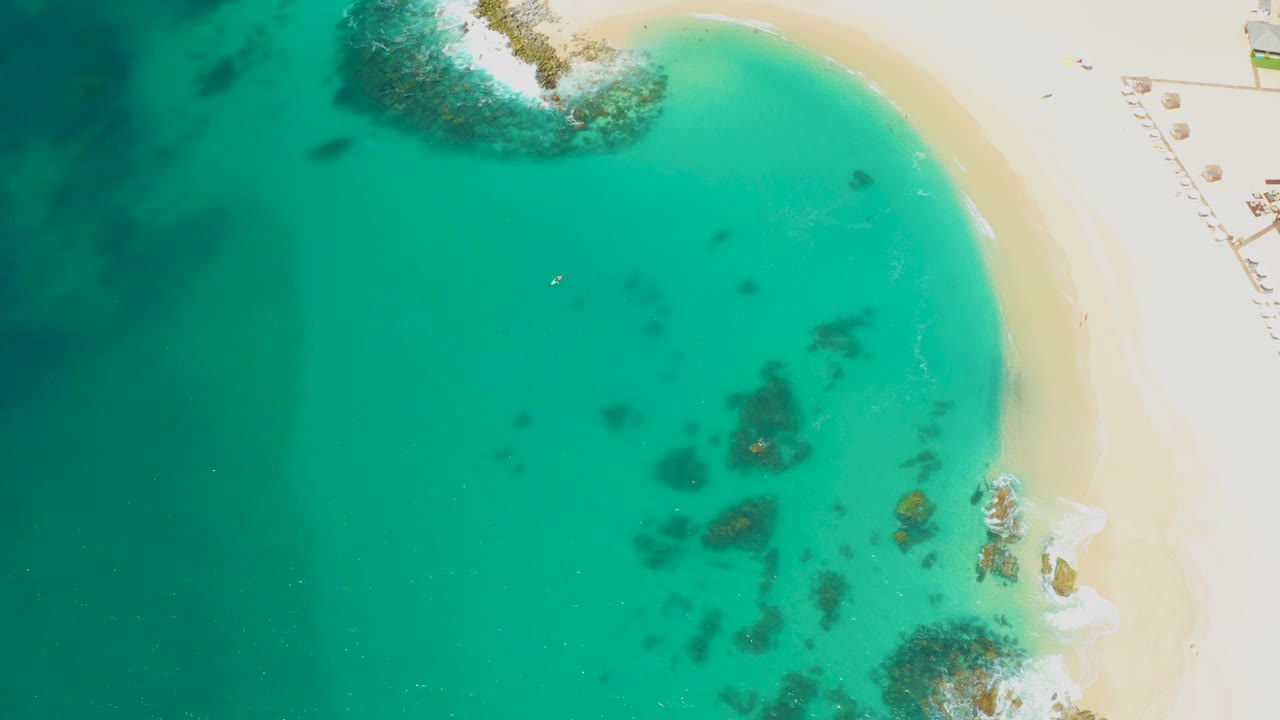 Aerial frame over Dreams Los Cabos water with the shoreline curving along the right, underwater rocks clustered near the lower right, white umbrellas on sand, and a tiny distant swimmer-like speck in the water.
