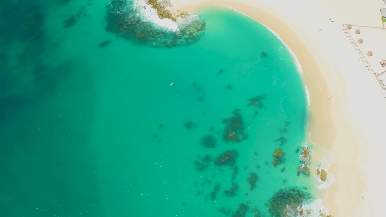 Top-down view of a curved sandy beach beside clear turquoise water.