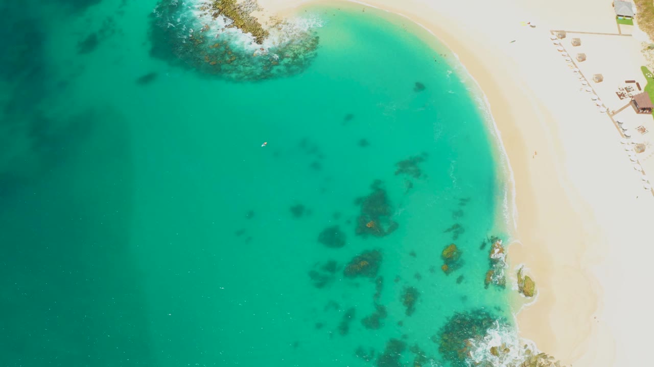 At 0:01, the Dreams Los Cabos shoreline remains on the right while darker underwater rock patches sit near the center of the turquoise water, with no recognizable people visible.