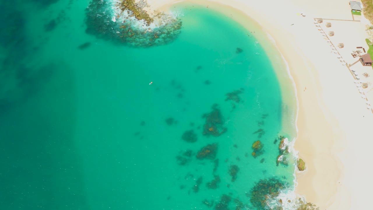 Aerial shoreline frame with green-blue water, sandy beach, and small surf breaks.