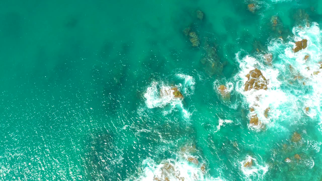 Aerial frame from Dreams Los Cabos with open turquoise water on the left, a bright foam patch near center, larger rocks and breaking surf on the right, and no visible people.