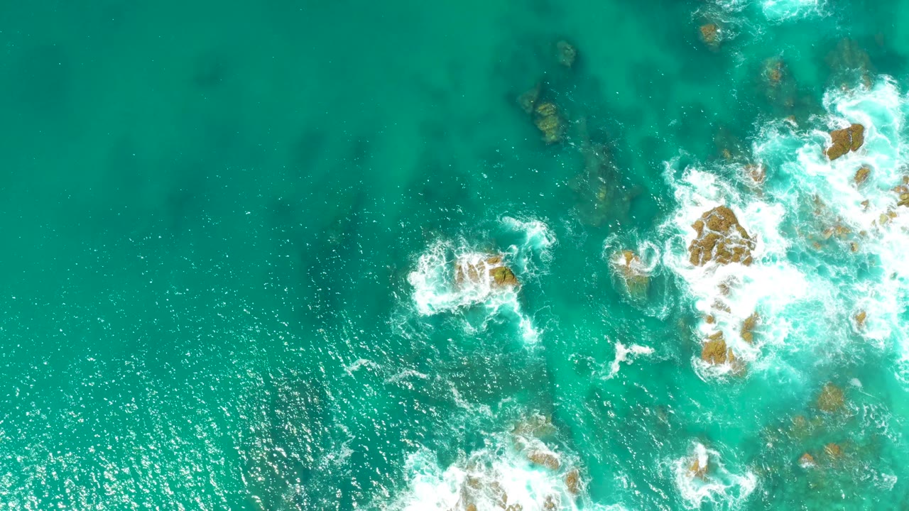 Top-down Dreams Los Cabos water frame where the rocky outcrop and white surf sit farther to the right, with rippled turquoise water and darker underwater patches across the left side, no visible people.