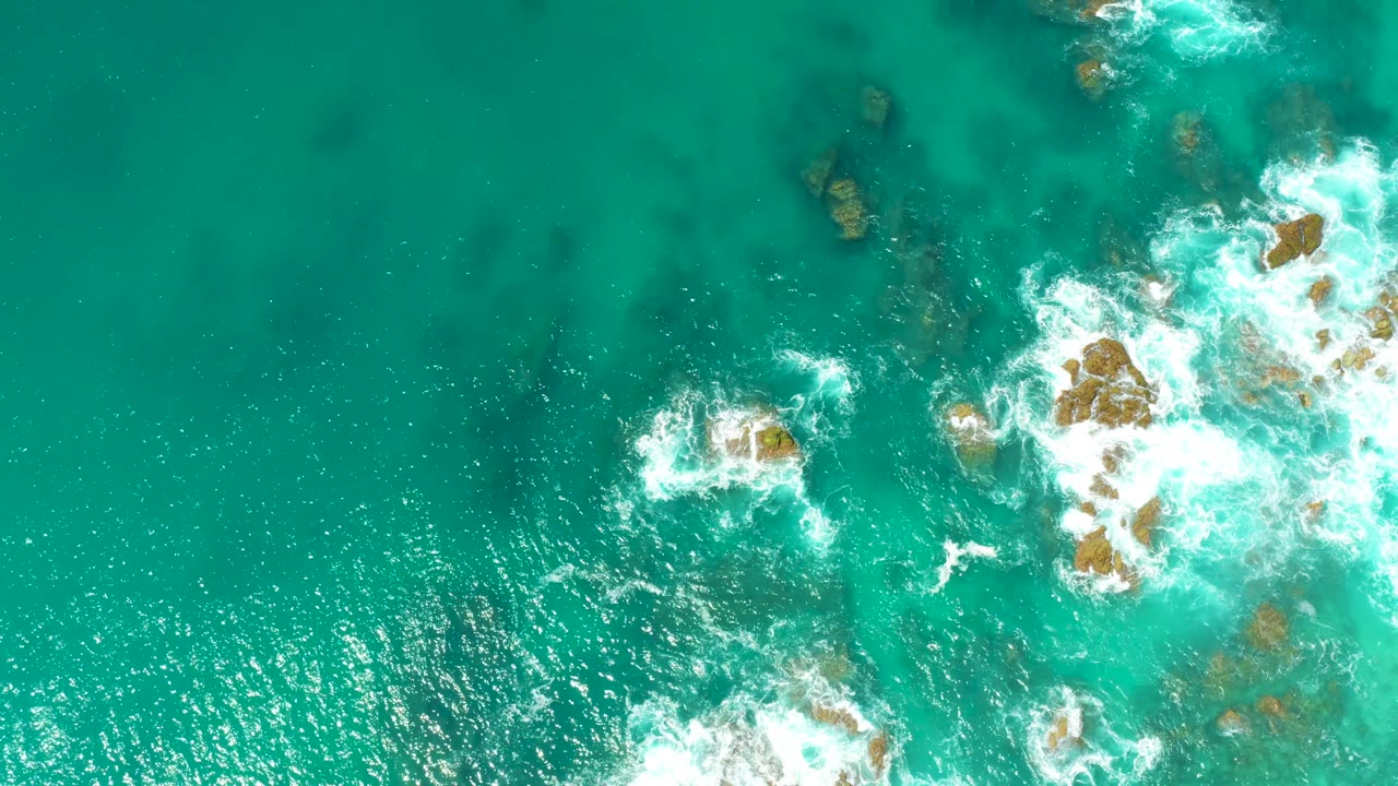 Dreams Los Cabos aerial view with a small foamy break near center-left, a larger cluster of rocks and whitewater on the right, bright green-blue water below, and no visible people.