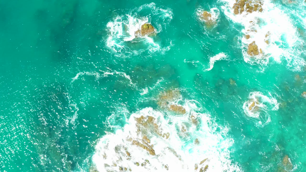Overhead view at Dreams Los Cabos showing turquoise water, white foam breaking around scattered rocks, and no visible people.