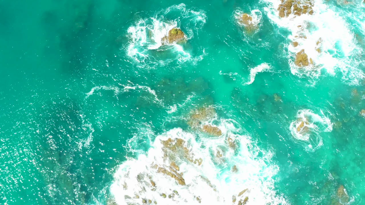 At 0:01, white surf expands over tan rocks beneath clear turquoise water at Dreams Los Cabos, with no visible people.