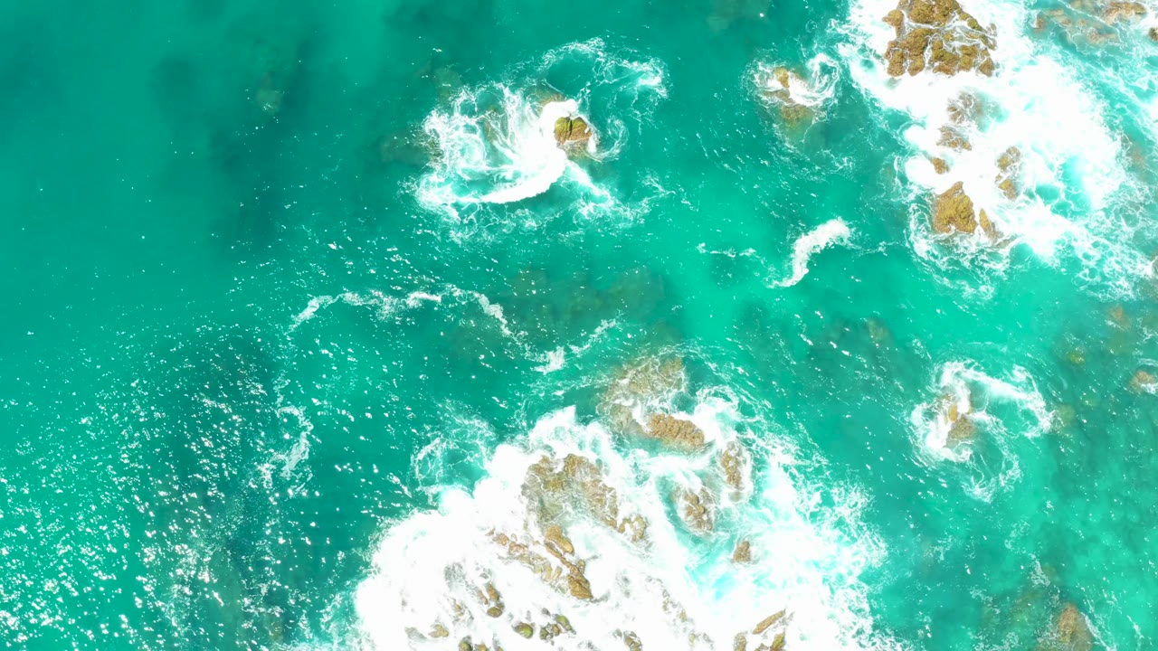 At 0:02, the overhead frame shows brighter foam crossing shallow rocky water near Dreams Los Cabos, with no visible people.