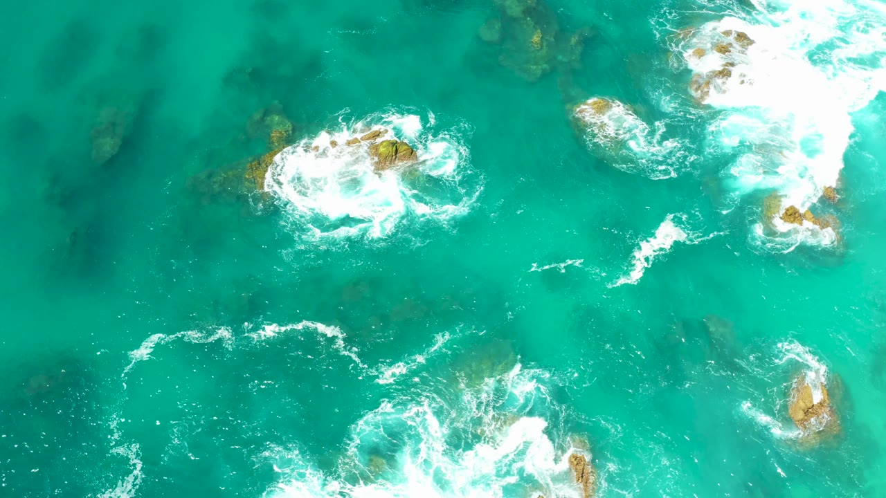 Overhead frame from Dreams Los Cabos showing foamy white water curling around scattered rocky outcrops beneath green-blue ocean, with no visible people.
