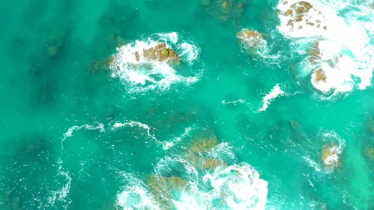 Overhead view at Dreams Los Cabos with a foamy rock near the upper center, a larger bright surf patch to the right, darker submerged rocks, and no visible people.