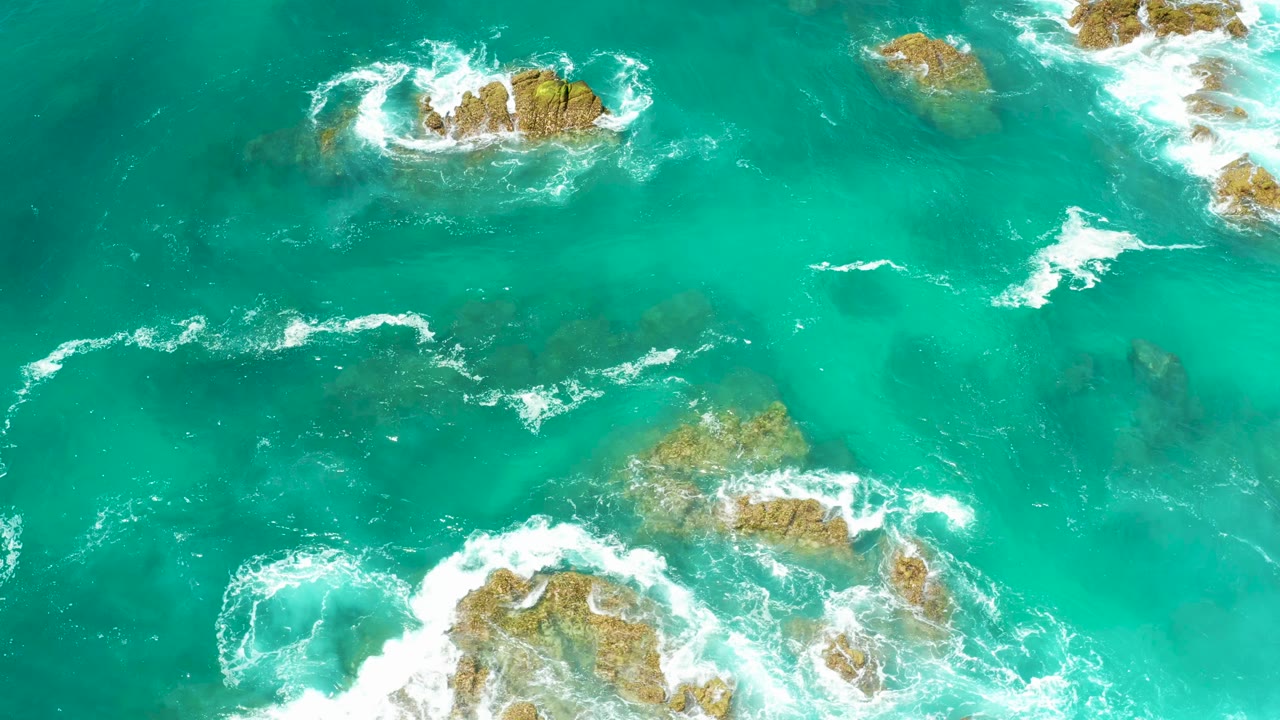 Aerial frame from Dreams Los Cabos with clear blue-green water, submerged rock shapes, and white surf lines drifting between exposed rocks, with no visible people.