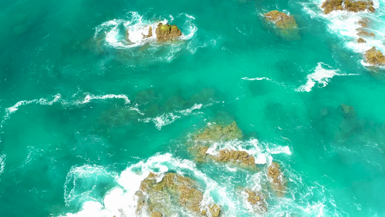 Overhead Los Cabos ocean detail with a bright foam break around upper and lower rocky outcrops, plus darker submerged patches beneath the water, with no visible people.